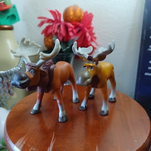 May include: Two plastic moose figurines standing side by side. The left moose is a darker brown with light gray antlers and hooves. The right moose is a lighter golden brown with similar light gray antlers and hooves. Both have large noses.