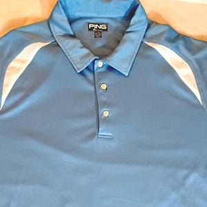 Könnte beinhalten: Hellblaues Poloshirt mit weißen Schulterakzenten. Das Shirt hat einen Kragen und drei Knöpfe. Der Markenname "PING" ist auf dem Kragen sichtbar.