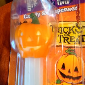 May include: Orange plastic Halloween-themed candy dispenser shaped like a jack-o'-lantern. The dispenser has a green stem and a smiling face. The packaging reads "Trick or Treat".