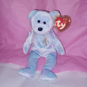 Puede incluir: Un oso de peluche azul claro con nariz y ojos negros, y un pequeño diseño de árbol bordado en el pecho. El oso tiene una etiqueta roja en forma de corazón sujeta a su oreja.