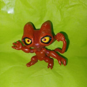 Könnte beinhalten: Kleine, rotbraune Alien-Figur mit großen gelben Augen und einer detaillierten, strukturierten Oberfläche. Die Figur hat ein einzigartiges, stilisiertes Design mit markanten Ohren und einem gebogenen Anhang. Das Spielzeug ist mit ausgestreckten Armen posiert, was ihm ein dynamisches Aussehen verleiht.