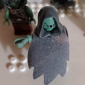 May include: A Lego minifigure of a green-skinned character wearing a gray hooded cloak. The figure has a detailed face and green hands. Another similar figure is visible in the background. The figures are on a white surface.