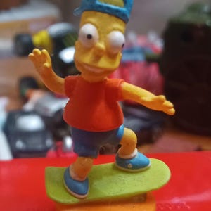 May include: A yellow cartoon character figurine wearing a blue cap, red t-shirt, and blue shorts, standing on a lime green skateboard. The figure is mounted on a round orange base, with both arms extended outward in a balanced pose.