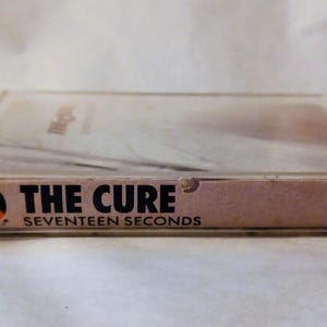 May include: A vintage cassette tape of The Cure's album "Seventeen Seconds." The cassette case is clear plastic, and the label is a light brown with black text. The Cure's logo is visible on the left side of the label.