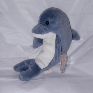 Puede incluir: Un peluche de delfín en tonos azules y blancos. El delfín tiene un cuerpo redondeado, un hocico prominente y ojos pequeños y negros. Tiene una barriga blanca y una etiqueta adjunta.