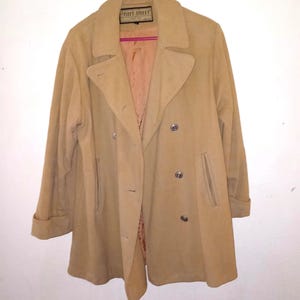 May include: A tan double-breasted coat with a wide collar, long sleeves with cuffs, and two vertical slit pockets. The coat features silver-toned buttons and a label at the neckline that reads Fleet Street Petite. The interior is lined in a lighter tan fabric.