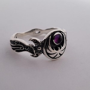 May include: A silver ring with an intricate design, featuring a central purple gemstone. The ring has a detailed, swirling pattern on the band and around the gemstone setting. The gemstone is round and set in a decorative bezel.
