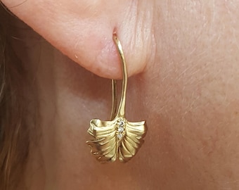 Puede incluir: Pendientes dorados con diseño de hoja. Cada pendiente presenta un gancho curvo y una forma de hoja texturizada y detallada con pequeños acentos brillantes. Los pendientes se muestran en una oreja.