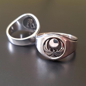 May include: Two silver rings with a detailed emblem design. One ring is in focus, displaying a circular emblem with a crescent moon and stylized bird. The other ring is out of focus, showing the inside of the ring.