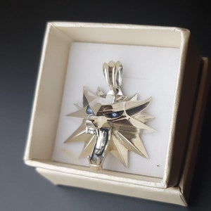 Puede incluir: Colgante de plata en una caja de regalo. El colgante presenta un diseño de cabeza de lobo con ojos de piedra azul y un marco en forma de estrella. El colgante se muestra en una caja beige con interior blanco, sobre un fondo oscuro.