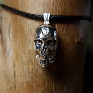 May include: A silver-colored pendant in the shape of a skull, with red accents in the eye sockets, hangs from a black braided cord. The skull pendant has detailed features and is displayed against a wooden background.