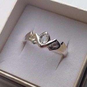 Puede incluir: Un anillo de plata con un diseño único, con tres piedras preciosas transparentes engastadas en un patrón geométrico. El anillo se presenta en una pequeña caja de joyería beige abierta, creando una exhibición limpia y elegante.
