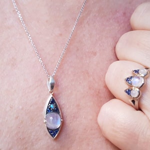 May include: A silver necklace with a teardrop-shaped pendant featuring a large, round, light-colored stone, surrounded by smaller blue stones. A matching ring with three similar stones is also visible.