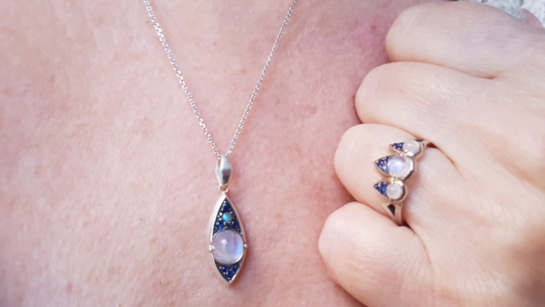 May include: A silver necklace with a teardrop-shaped pendant featuring a large, round, light-colored stone, surrounded by smaller blue stones. A matching ring with three similar stones is also visible.