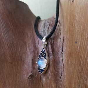 May include: A silver pendant necklace with a teardrop-shaped design. The pendant features a large, round, iridescent moonstone, surrounded by small blue and black stones. The necklace has a black cord.