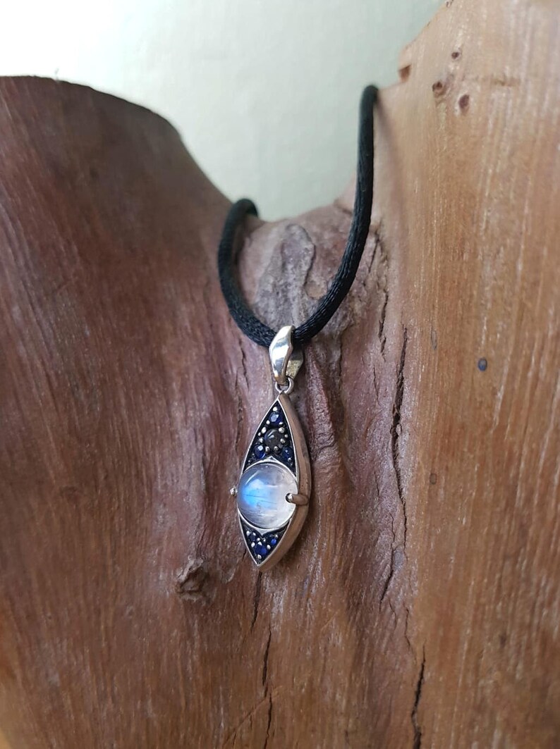 May include: A silver pendant necklace with a teardrop-shaped design. The pendant features a large, round, iridescent moonstone, surrounded by small blue and black stones. The necklace has a black cord.