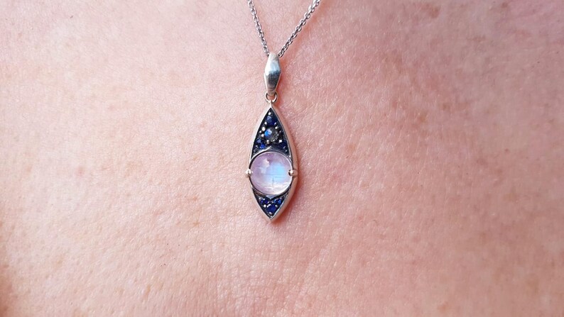 May include: A silver pendant necklace featuring a teardrop-shaped design. The pendant has a central, round, iridescent stone, surrounded by small blue stones. The necklace is displayed against a skin tone background.
