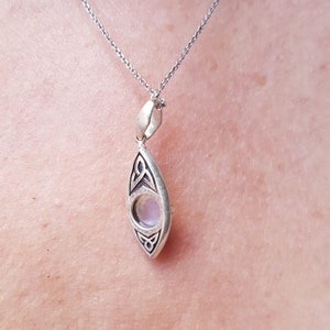 May include: Silver pendant necklace with a teardrop-shaped pendant. The pendant features a light-colored, round stone set within a silver frame with Celtic knot designs. The necklace chain is also silver.