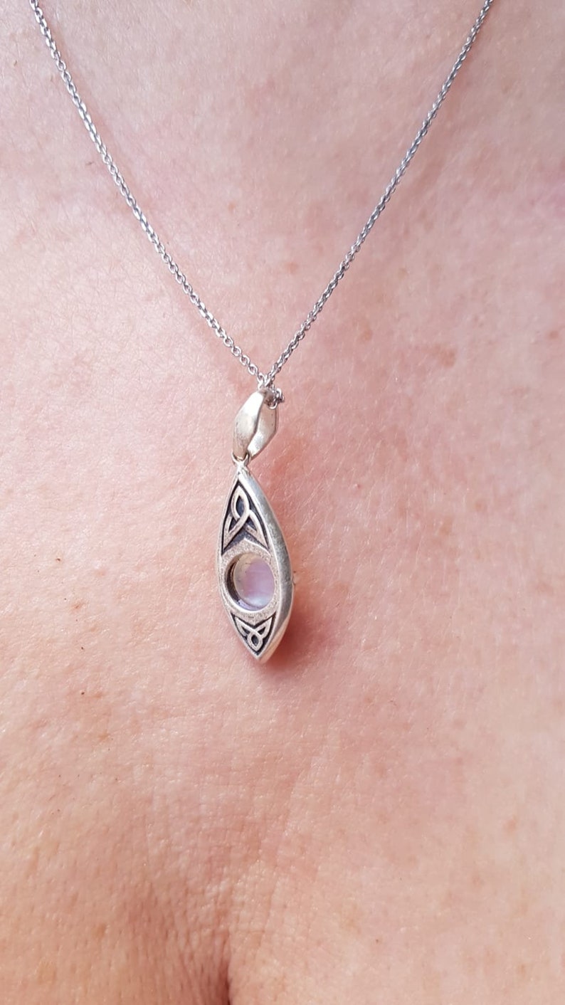 May include: Silver pendant necklace with a teardrop-shaped pendant. The pendant features a light-colored, round stone set within a silver frame with Celtic knot designs. The necklace chain is also silver.
