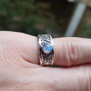 Può includere: Un anello d'argento con un motivo a spirale ornato e una pietra di luna iridescente, rotonda e centrale. L'anello è mostrato su un dito, con lo sfondo sfocato.