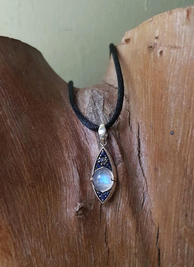 May include: A silver pendant necklace with a teardrop-shaped design. The pendant features a large, round, iridescent moonstone in the center, surrounded by small, dark blue gemstones. The necklace has a black cord.