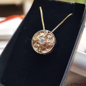 Op de afbeelding: Een goudkleurige ronde hanger aan een ketting met een ingewikkeld opengewerkt filigraanontwerp en een enkele heldere, rondgeslepen steen in het midden. De hanger hangt aan een fijne goudkleurige ketting en wordt gepresenteerd in een zwarte doos.
