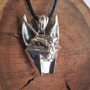 Puede incluir: Un collar con colgante de plata que representa una cabeza de gato estilizada con orejas puntiagudas y ojos de piedras preciosas rojas. El colgante cuelga de un cordón negro. La cara del gato tiene detalles geométricos y está muy pulida.