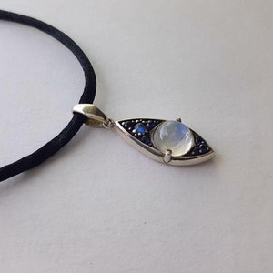 May include: Silver pendant necklace featuring an eye-shaped design. The pendant has a central moonstone, surrounded by small blue gemstones. The necklace has a black cord.