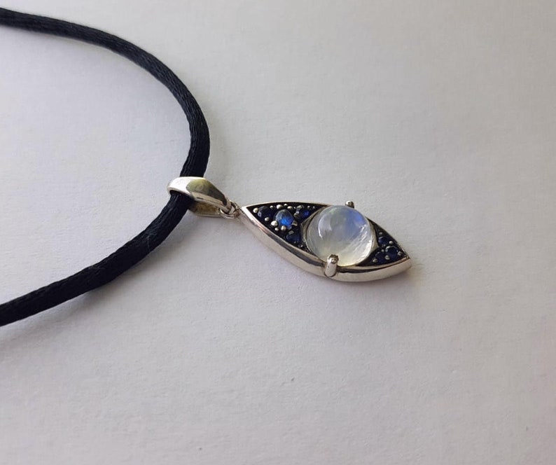 May include: Silver pendant necklace featuring an eye-shaped design. The pendant has a central moonstone, surrounded by small blue gemstones. The necklace has a black cord.