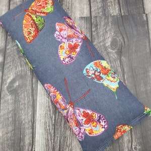 May include: A gray fabric eye pillow with a colorful butterfly pattern. The pillow is filled with flax seeds and lavender.
