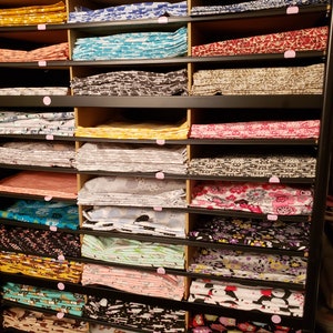 May include: A black metal shelving unit with multiple shelves filled with stacks of colourful fabric. The fabric is folded and arranged neatly on the shelves. The fabric has various patterns and designs, including floral, geometric, and animal prints.