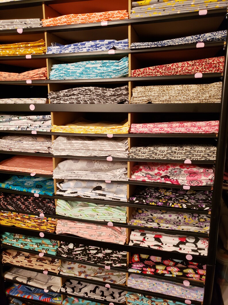 May include: A black metal shelving unit with multiple shelves filled with stacks of colourful fabric. The fabric is folded and arranged neatly on the shelves. The fabric has various patterns and designs, including floral, geometric, and animal prints.