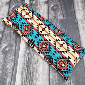 May include: A rectangular turquoise, red, black, and white patterned eye pillow. The pillow has a geometric design with a repeating pattern of triangles and squares.