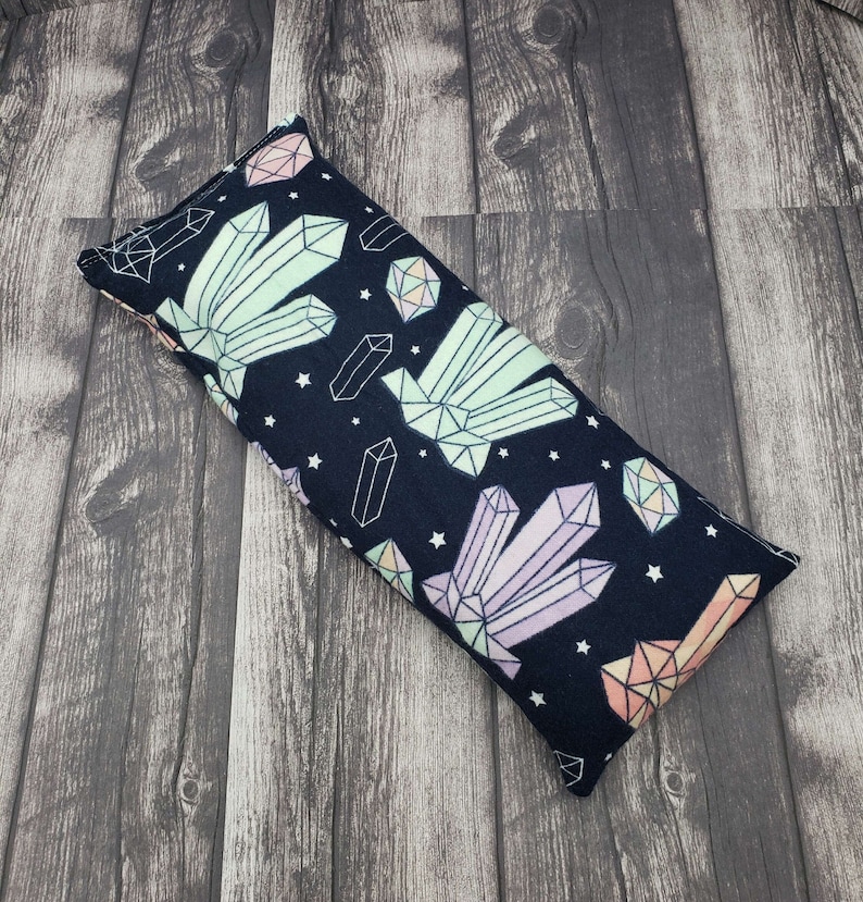 May include: A black fabric eye pillow with a colourful geometric crystal pattern. The pillow is filled with flax seeds and lavender.