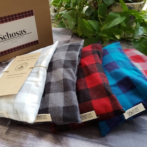 May include: Five different plaid fabric rice and flaxseed heating pads. The pads are arranged in a row from left to right, with the white pad on the left and the teal pad on the right. The pads are all tied with twine and have a label that says "Sehosas" on them.