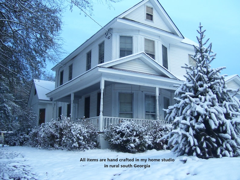 Puede incluir: Una casa blanca con un porche y un &aacute;rbol de hoja perenne cubierto de nieve en frente. La casa tiene una valla blanca y un c&eacute;sped cubierto de nieve. El texto "All items are hand crafted in my home studio in rural south Georgia" es visible en la parte inferior de la imagen.