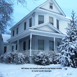 May include: A white house with a porch and a snow-covered evergreen tree in front of it. The house has a white picket fence and a snow-covered lawn. The text "All items are hand crafted in my home studio in rural south Georgia" is visible at the bottom of the image.