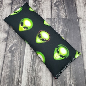 May include: A black fabric eye pillow with a repeating pattern of green alien faces wearing sunglasses. The pillow is rectangular and has a seam around the edges.