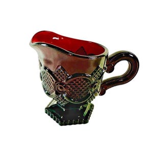 May include: A dark red iridescent glass creamer featuring a textured diamond pattern on the exterior, a curved handle, and a scalloped base. The interior of the vessel is a deep, solid red, while the outer surface displays a metallic sheen.