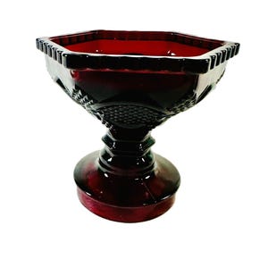 May include: A deep red glass pedestal bowl featuring an octagonal rim with a scalloped edge. The exterior of the bowl displays a textured diamond pattern across its sides, supported by a thick, multi-tiered circular base made of matching red glass.