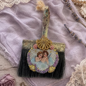 May include: A decorative paintbrush featuring a weathered handle and black bristles. The brush head is adorned with a gold-toned shell ornament, two small pink roses, and a central circular image of two cherubs surrounded by purple flowers.