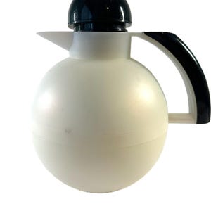 May include: A spherical thermal carafe featuring a matte white body, a glossy black dome-shaped lid, and a black curved handle. The vessel includes a pointed spout extending from the upper section, designed for pouring liquids while maintaining temperature.