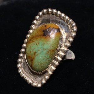 Pu&ograve; includere: Un anello d'argento con una pietra turchese verde e marrone incastonata in una lunetta a forma di conchiglia. L'anello &egrave; in argento sterling e ha una finitura testurizzata.