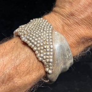 May include: A silver cuff bracelet with a textured design. The bracelet features a section covered in small, round beads and a smooth, curved section. The jewelry is worn on a wrist against a dark background.