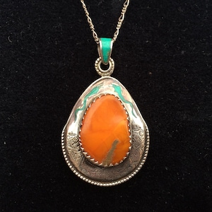 May include: Silver pendant necklace with an orange stone center. The pendant has a teardrop shape with turquoise accents and intricate silver detailing. The necklace chain is a delicate silver.