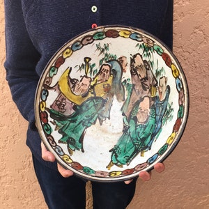 May include: A round, antique ceramic plate with a black rim and a colorful border. The plate features a hand-painted scene of figures in green, yellow, and brown robes. The plate is being held by a person wearing a navy blue sweater and dark blue jeans.