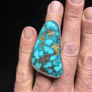 May include: A large turquoise ring with a silver setting. The turquoise stone is a vibrant blue-green color with brown and gold matrix patterns. The ring is set on a finger, showcasing the size and shape of the stone.
