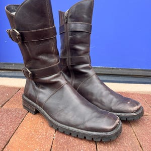 May include: A pair of brown leather boots with a mid-calf height. The boots feature a zipper, two buckle straps, and a lug sole. The boots are worn and show signs of use.
