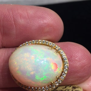 Op de afbeelding: Een gouden ring met een grote ovale opaal-edelsteen omringd door een halo van kleine diamanten. De opaal heeft een witte basis met flitsen van groen, rood en geel.