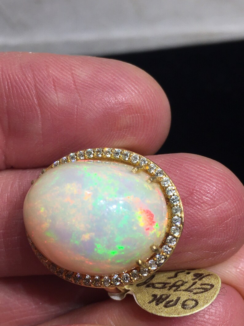 Op de afbeelding: Een gouden ring met een grote ovale opaal-edelsteen omringd door een halo van kleine diamanten. De opaal heeft een witte basis met flitsen van groen, rood en geel.